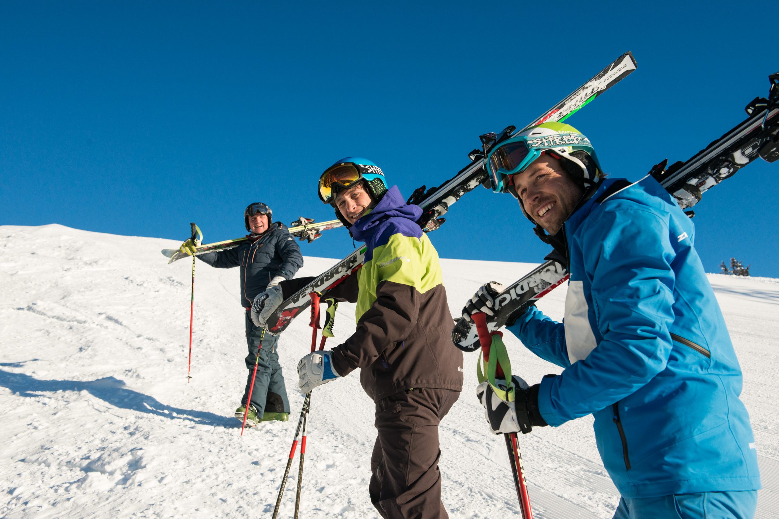 Skischule Grasgehren + Fischen drei Skifahrer laufen den Berg mit den Skiern auf der Schulter hinauf